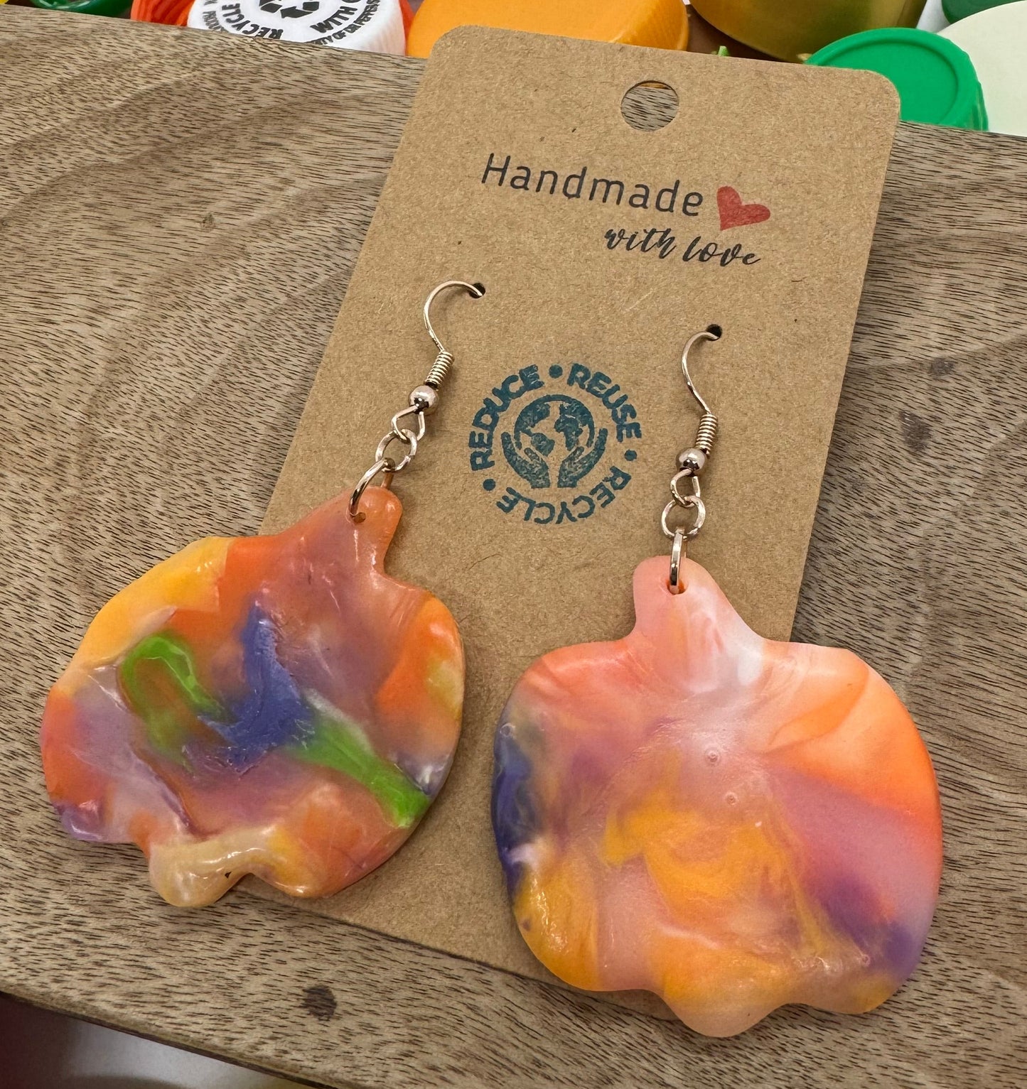 Colorful pumpkin shaped earrings on a wooden display card with a blurred background of colorful bottle caps.