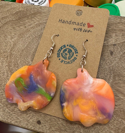 Colorful pumpkin shaped earrings on a wooden display card with a blurred background of colorful bottle caps.