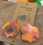 Colorful pumpkin shaped earrings on a wooden display card with a blurred background of colorful bottle caps.