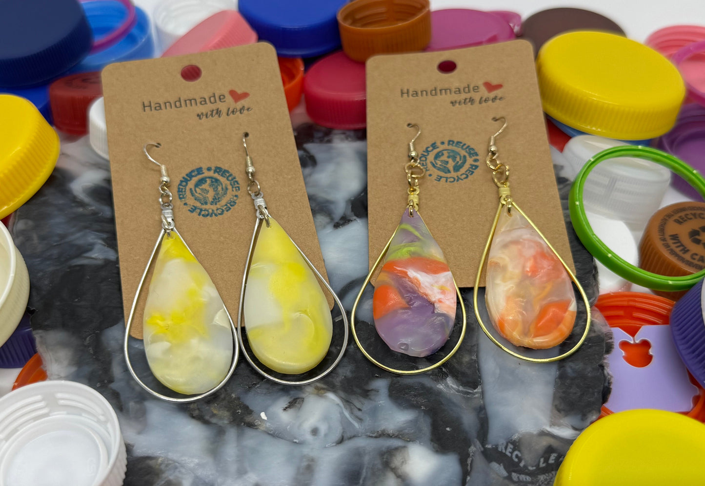 Two pairs of teardrop-shaped earrings on cards with colorful bottle caps in the background.