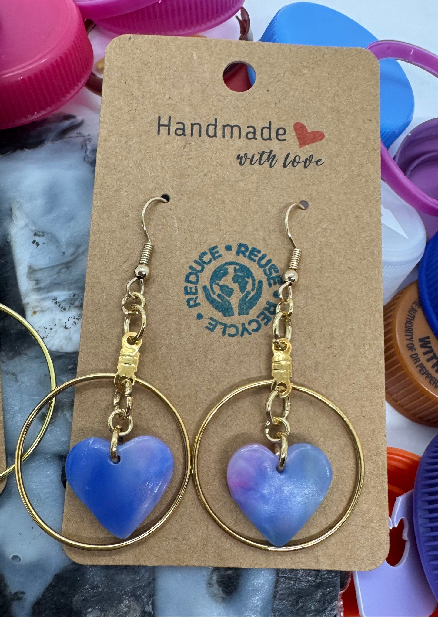 Gold hoop earrings with purple heart charms on a branded card, surrounded by colorful bottle caps.