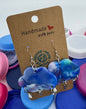 Blue and white beaded earrings on a 'Handmade' card with colorful bottle caps in the background.