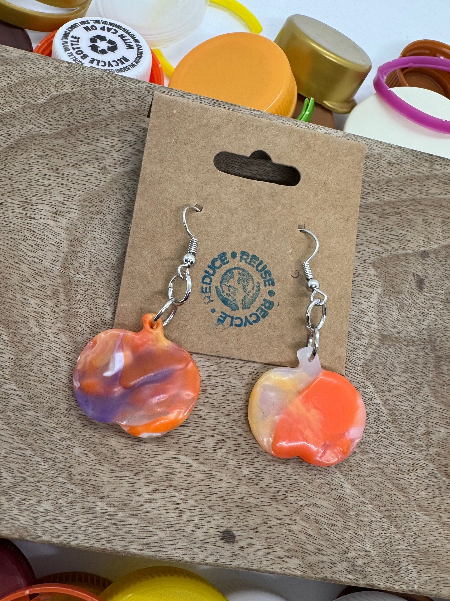 Colorful earrings on a cardboard backing with a wooden surface and colorful bottle caps in the background.