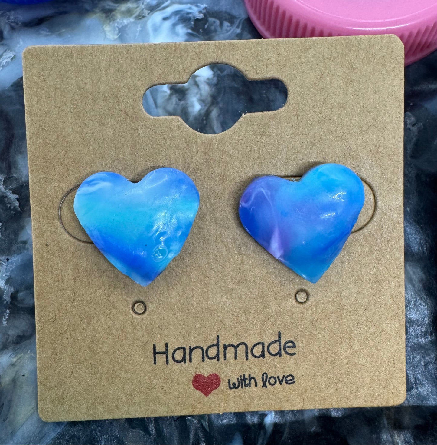 Heart-shaped blue earrings on a brown card with 'Handmade with Love' text, surrounded by plastic bottle caps.