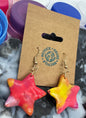 Star-shaped earrings on a marble surface with a cardboard holder and plastic lids.