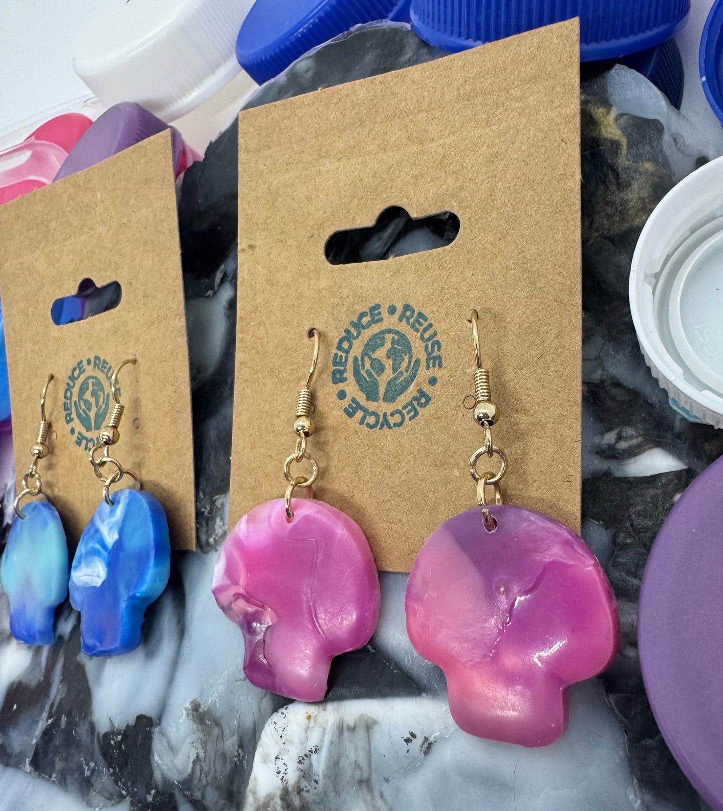 Pair of pink and blue earrings on a Reuse Recycle branded card with plastic bottle caps in the background.