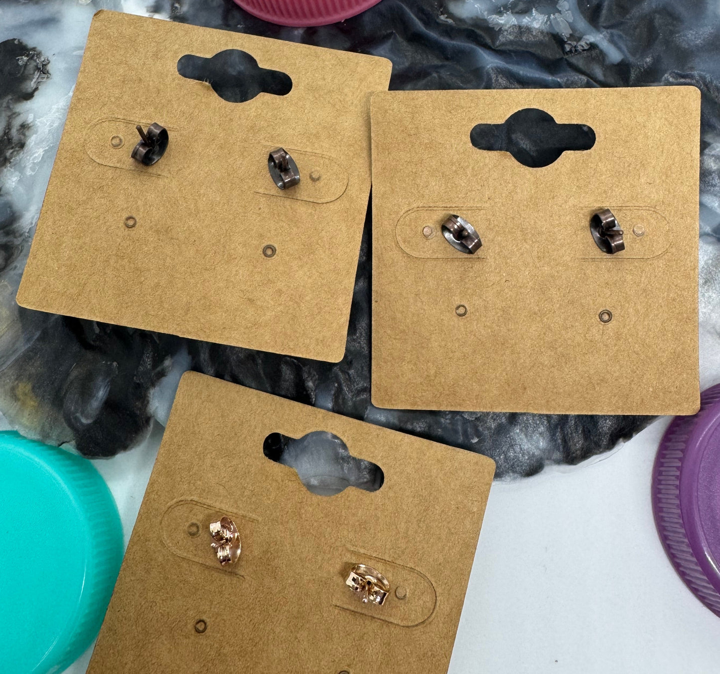 Three pairs of earrings on cardboard display cards with colorful bottle caps in the background.