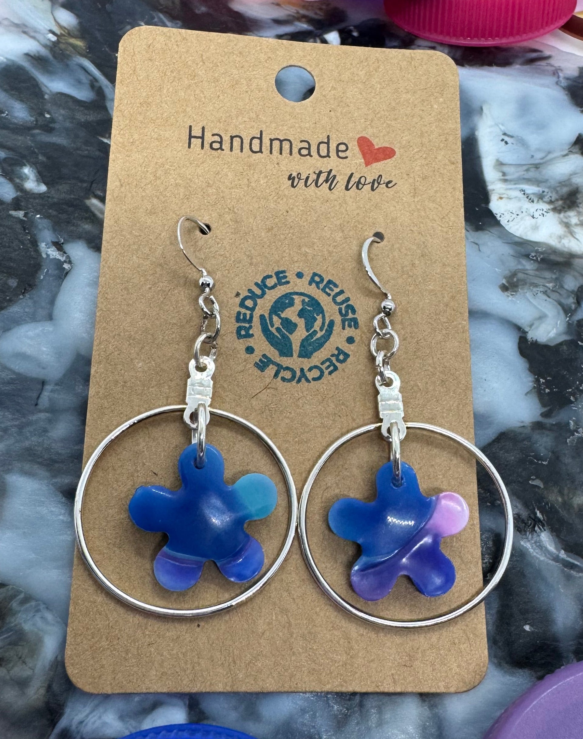 Pair of blue flower earrings on a cardboard card with 'Handmade' text, surrounded by colorful bottle caps.