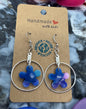 Pair of blue flower earrings on a cardboard card with 'Handmade' text, surrounded by colorful bottle caps.
