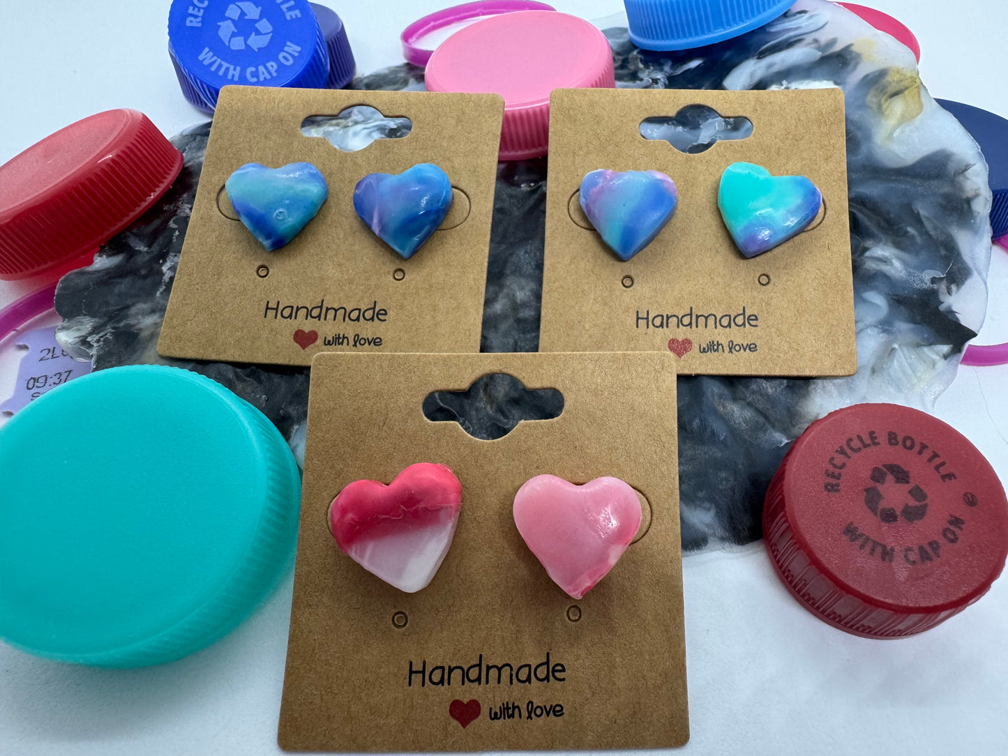 Heart-shaped earrings on a cardboard backing with 'Handmade' text, surrounded by plastic bottle caps.