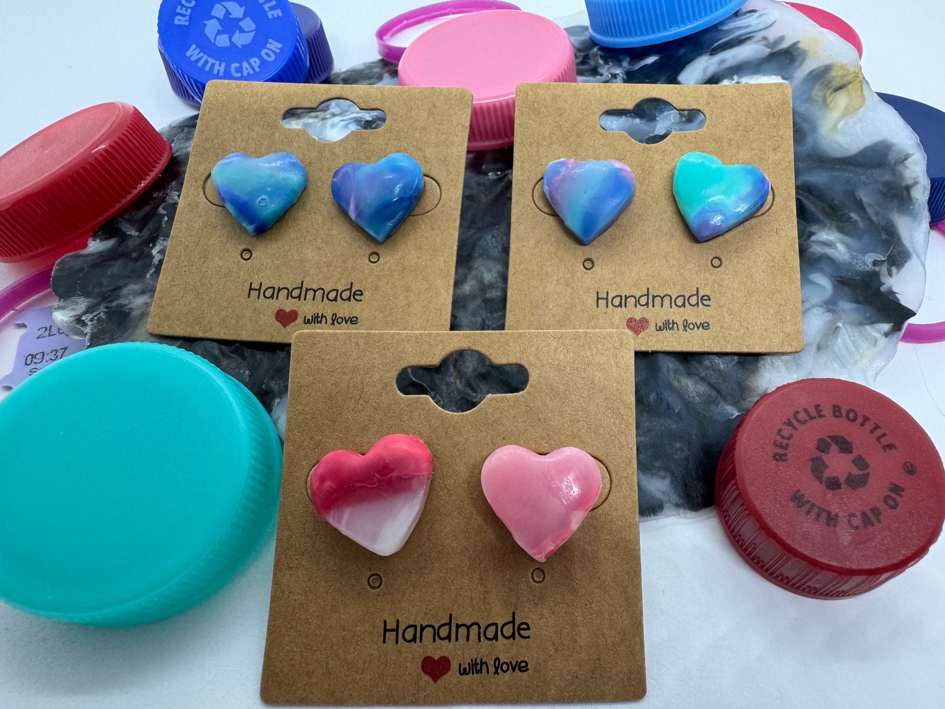 Heart-shaped earrings on a cardboard backing with 'Handmade' text, surrounded by plastic bottle caps.