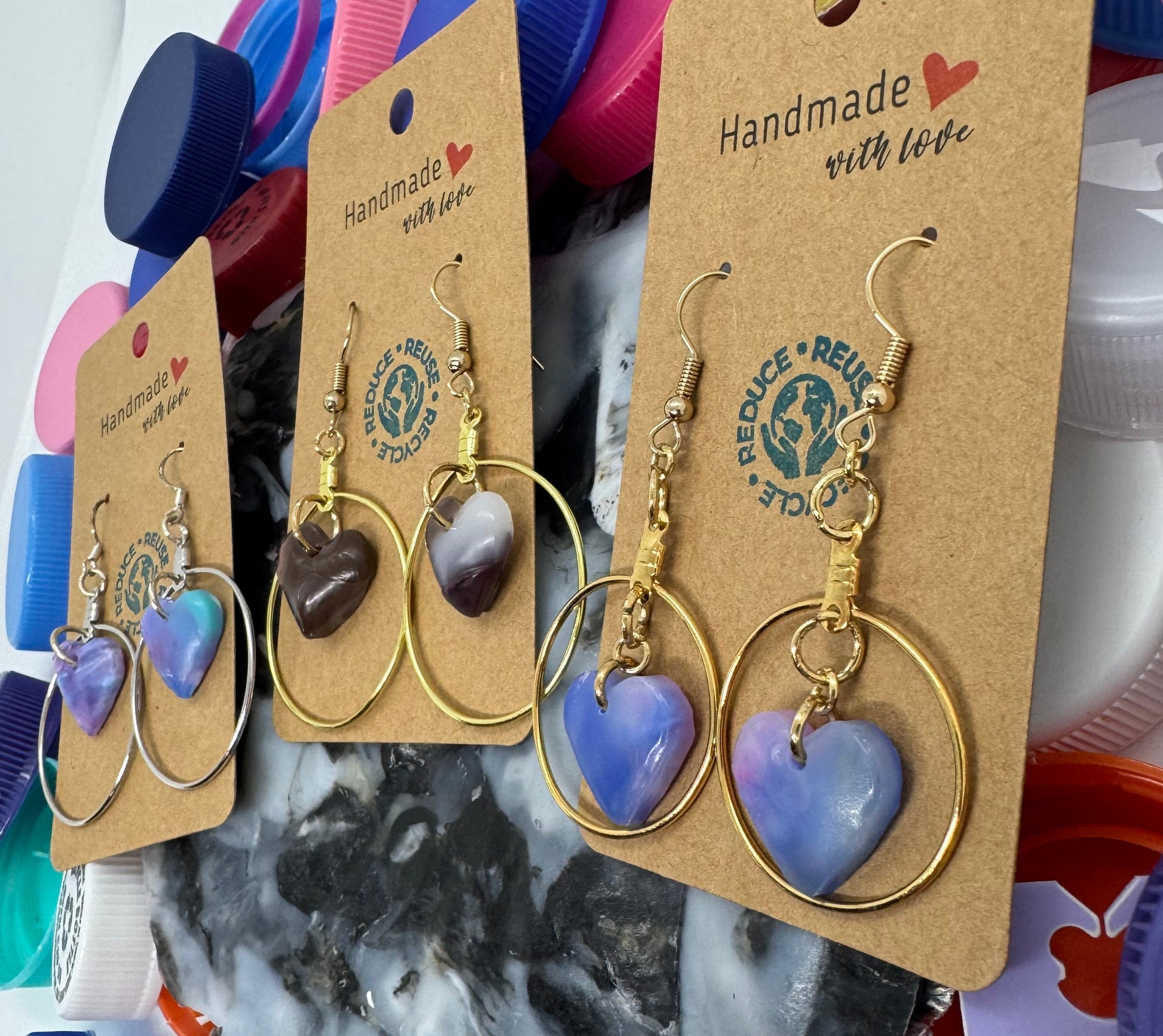 Gold heart-shaped earrings on a cardboard backing with colorful bottle caps in the background.