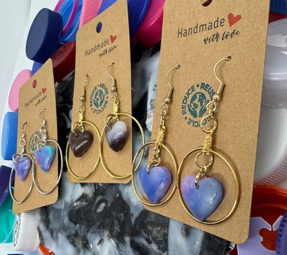 Gold heart-shaped earrings on a cardboard backing with colorful bottle caps in the background.