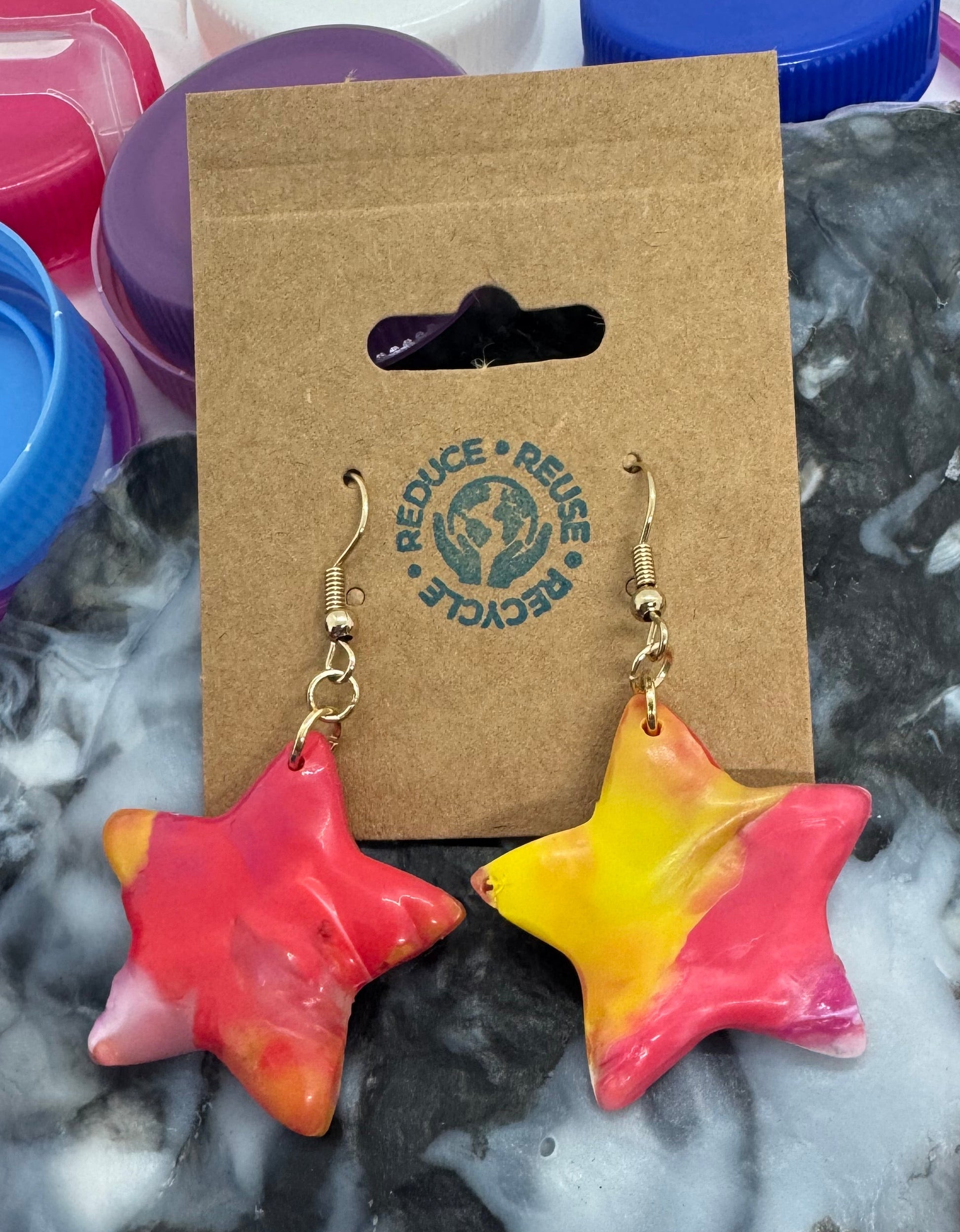 Colorful star-shaped earrings on a cardboard card with a brand logo, surrounded by plastic bottle caps.