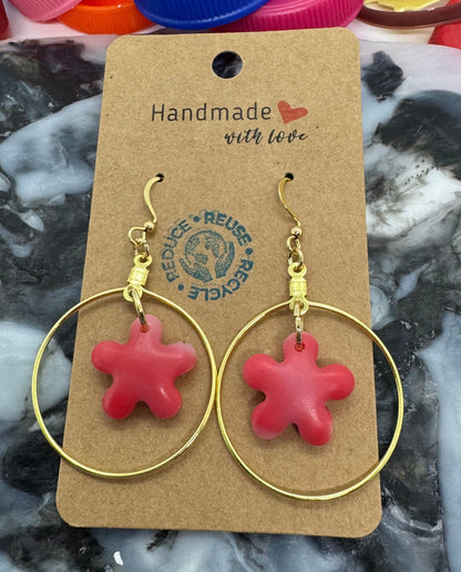 Red flower-shaped earrings on a gold hoop with a 'Handmade' label, placed on a textured surface with colorful bottle caps.
