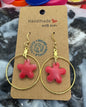 Red flower-shaped earrings on a gold hoop with a 'Handmade' label, placed on a textured surface with colorful bottle caps.