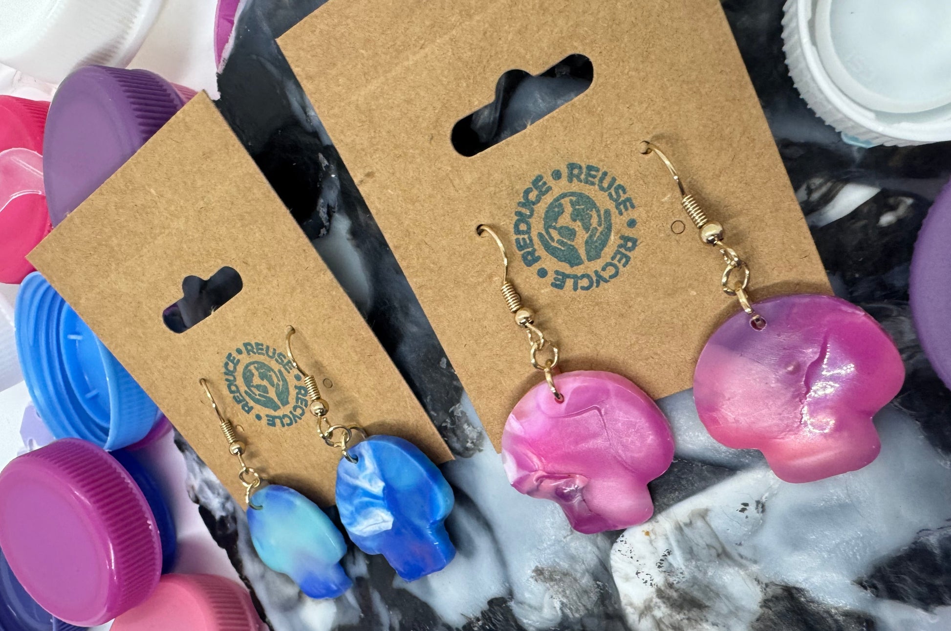 Pink and blue earrings on a cardboard display card with a visible brand logo, surrounded by plastic bottle caps.