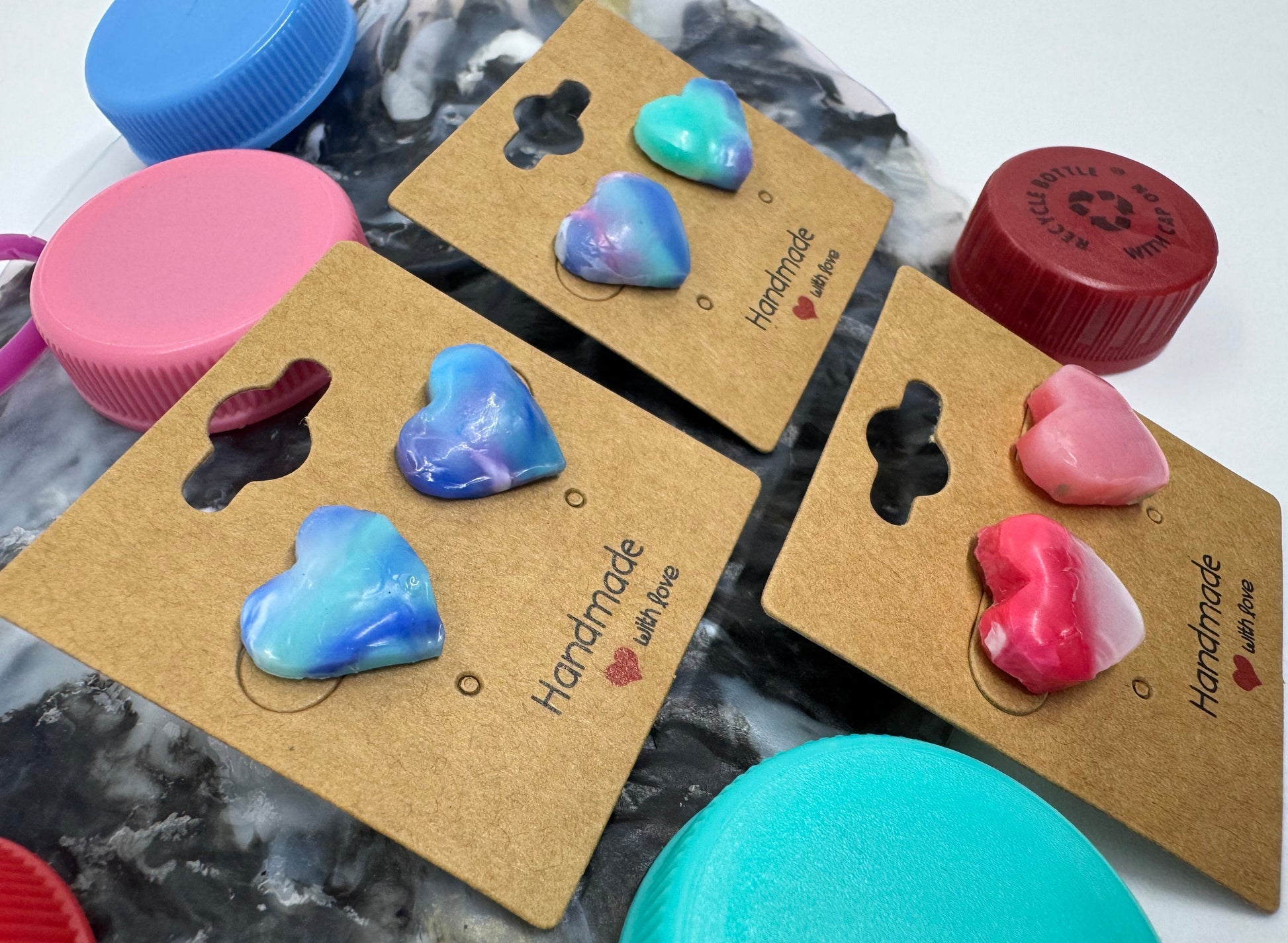 Heart-shaped earrings on brown cards with 'Handmade' text, surrounded by colorful bottle caps.