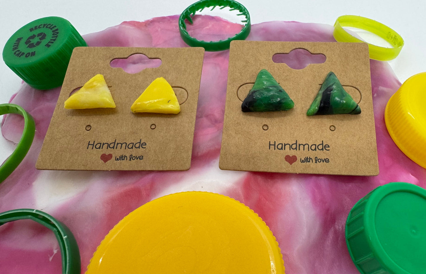 Two pairs of triangle-shaped earrings on cardboard cards with colorful plastic toys in the background.