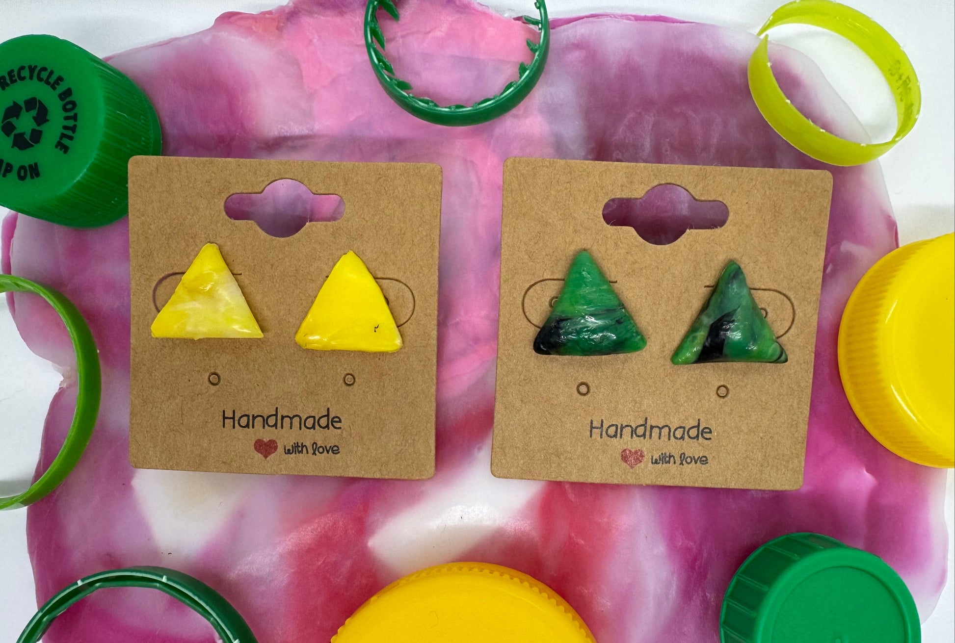 Two pairs of triangle-shaped earrings on cardboard cards with colorful bottle caps around.
