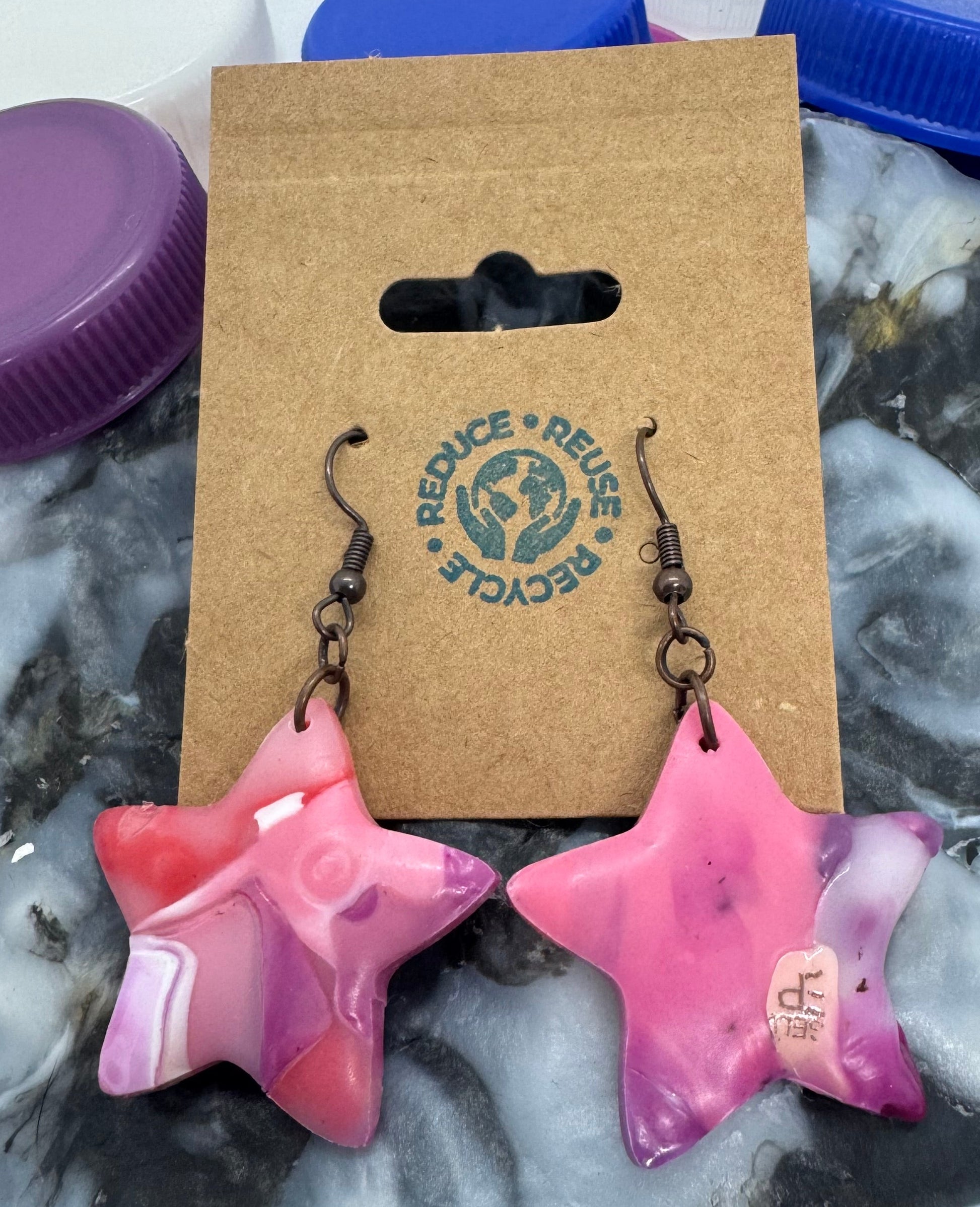 Pink star-shaped earrings on a card with a brand logo, placed on a textured surface.