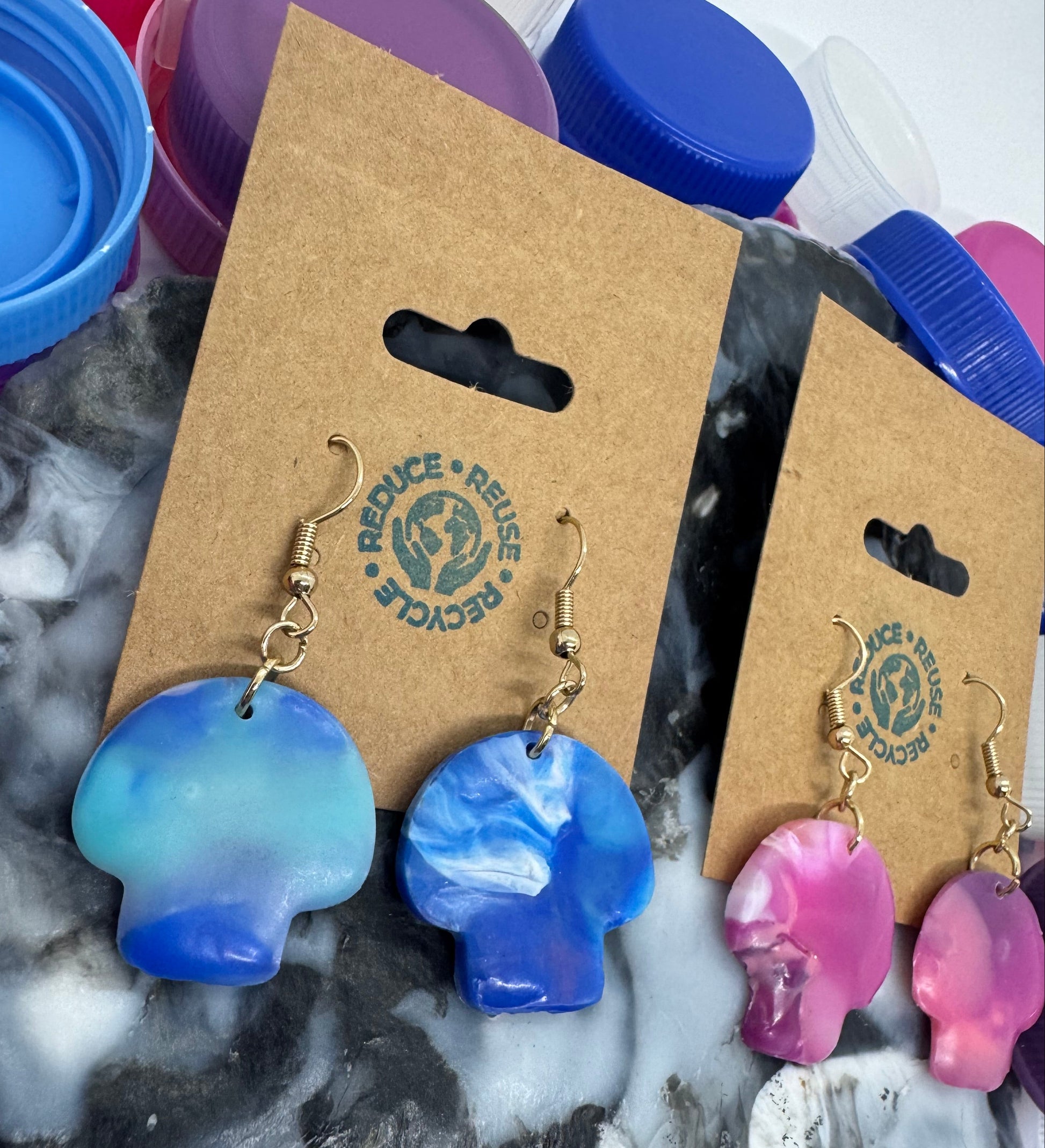 Blue and pink earrings on a cardboard display card with REUSE & RECYCLE branding, surrounded by plastic bottle caps.
