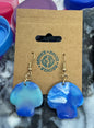 Blue earrings on a cardboard backing with colorful bottle caps in the background
