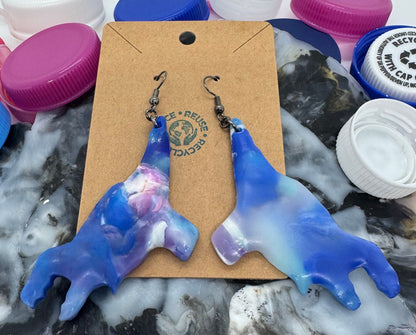 Blue and white ceramic dog-shaped earrings on a card with colorful bottle caps in the background.