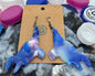 Blue and white ceramic dog-shaped earrings on a card with colorful bottle caps in the background.