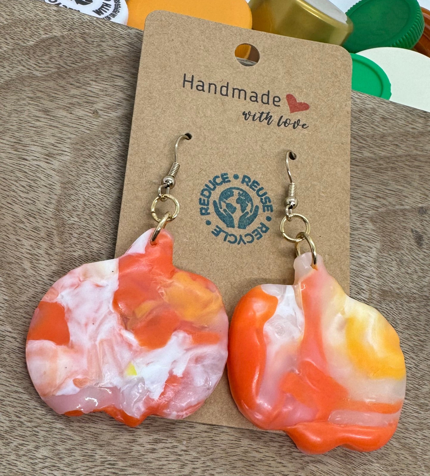Colorful earrings on a wooden stand with a 'Handmade' label, surrounded by colorful bottle caps.