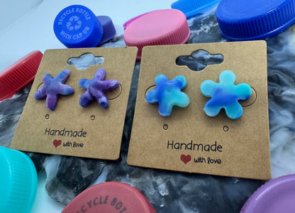 Pair of handmade flower-shaped earrings on cardboard cards with 'Handmade with love' text, surrounded by plastic bottle caps.