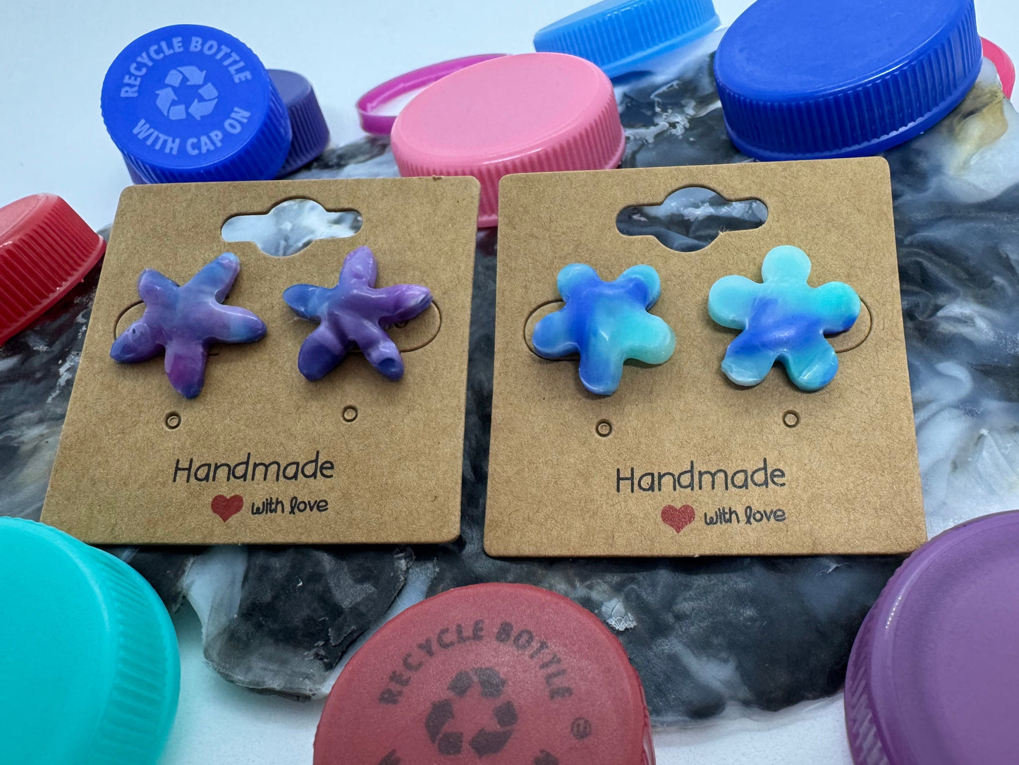 Pair of flower-shaped earrings on cardboard cards with colorful bottle caps in the background.